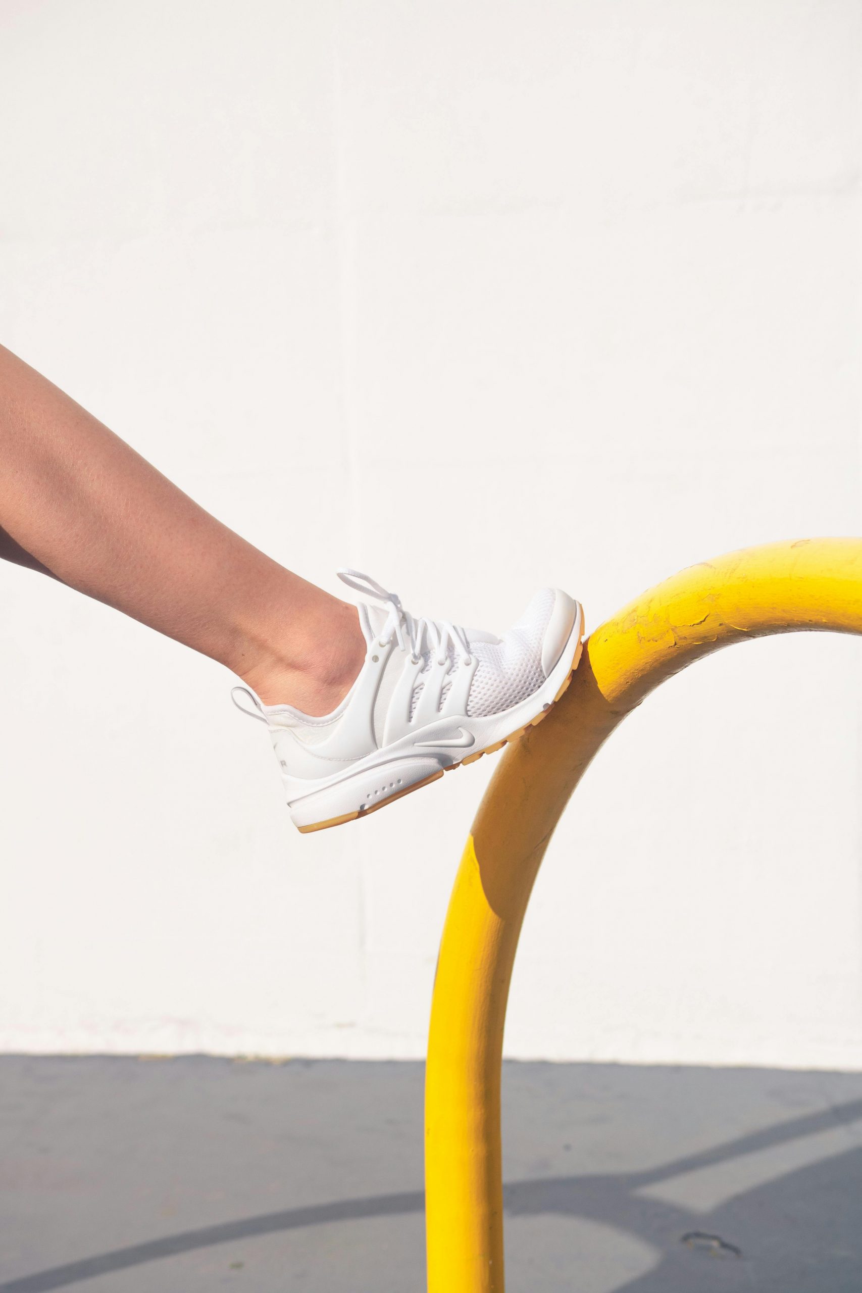 A person wearing stylish white sneakers resting foot on a curved yellow rail outdoors.