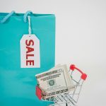Miniature shopping cart with cash next to a sale-tagged bag, illustrating shopping and savings.